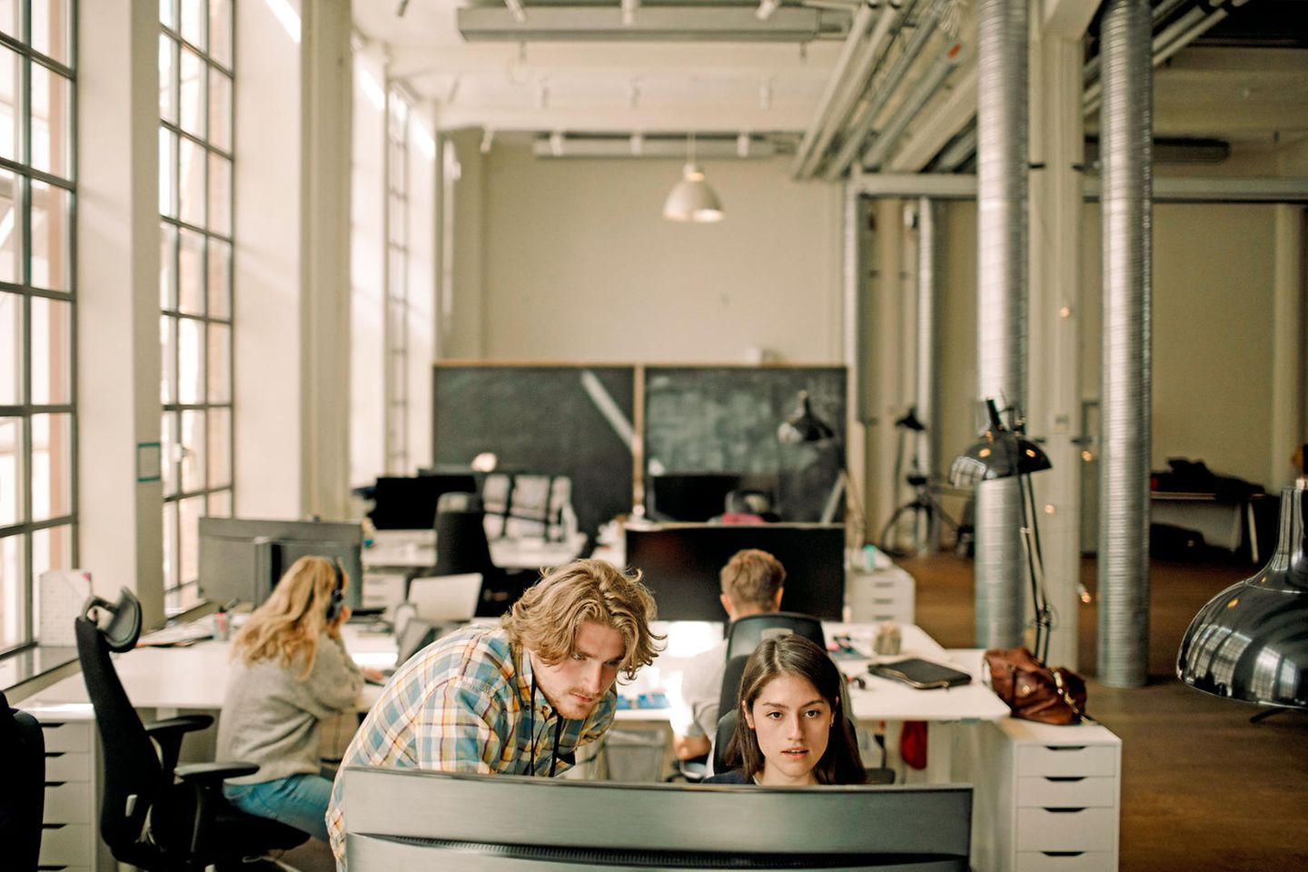Eine junge Frau sitzt in einem grossräumigen Büro vor ihrem Monitor und neben ihr steht ein junger Kollege helfend zu ihr