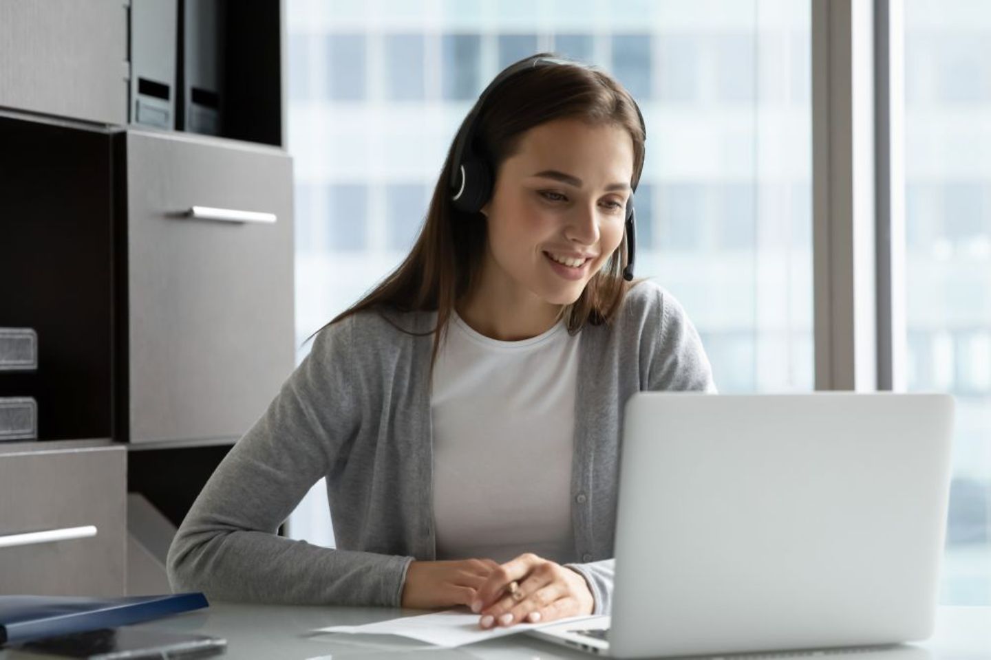 Junge Mitarbeiterin mit Headset, die online lernt und eine Schulung am Laptop im Büro verfolgt