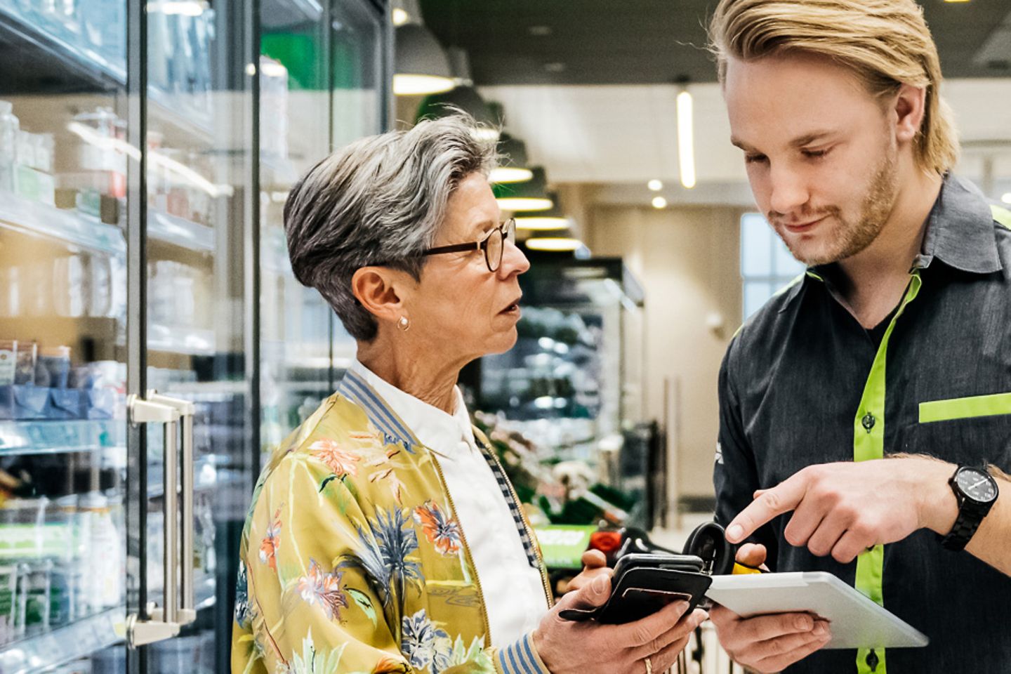 Junger Verkäufer und Marktleiterin stehen vor dem Kühlregal und gleichen Daten auf einem Tablet ab