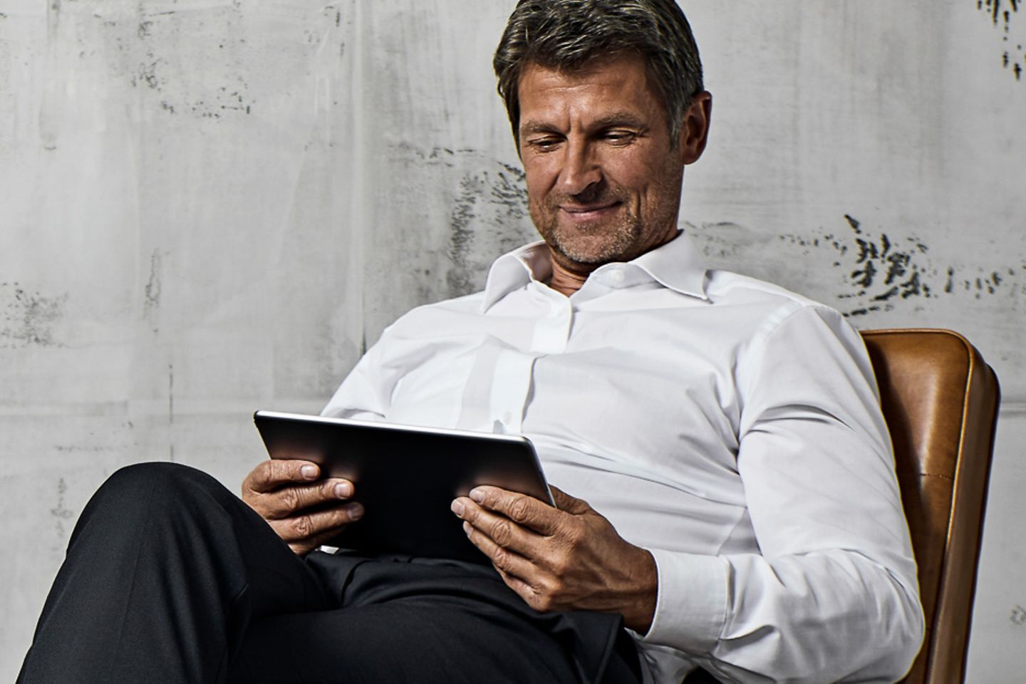 Businessman sits in leather armchair in front of concrete wall and reads on his tablet