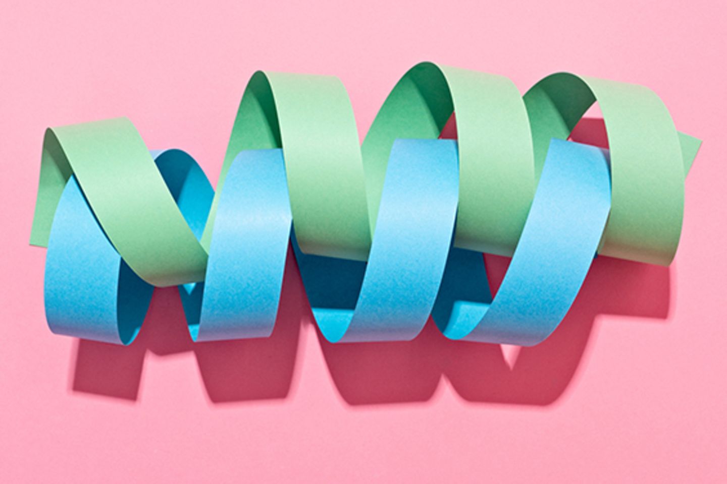 Grüne und blaue, ineinander gedrehte Papierspiralen auf rosa Untergrund. 