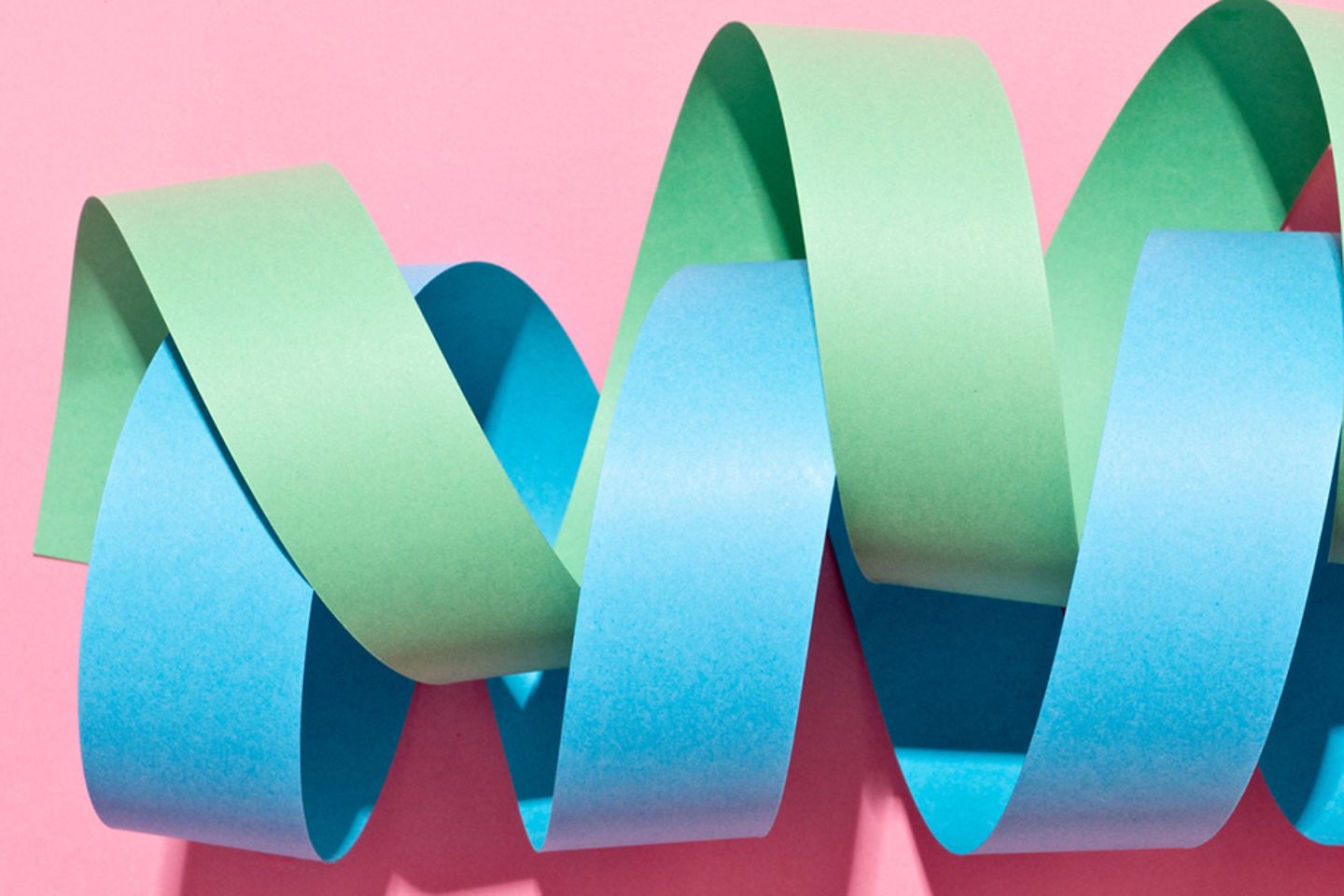 Grüne und blaue, ineinander gedrehte Papierspiralen auf rosa Untergrund. 