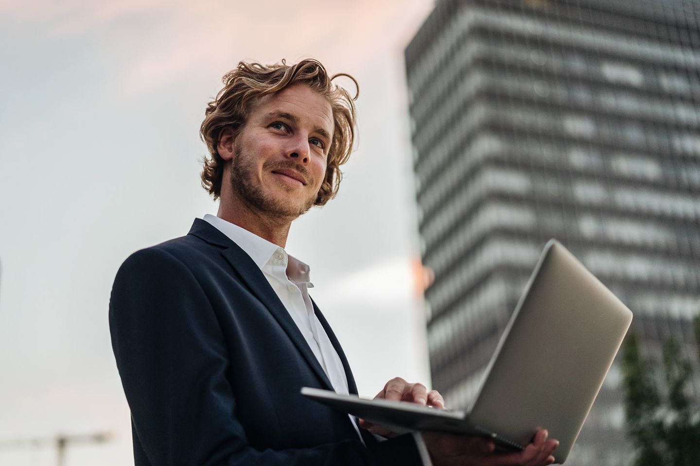 Un homme heureux se tient devant un bâtiment de bureaux avec un ordinateur ouvert dans les mains.