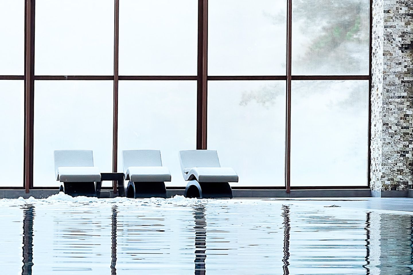 Chaises longues au bord d’une piscine couverte devant une immense baie vitrée.