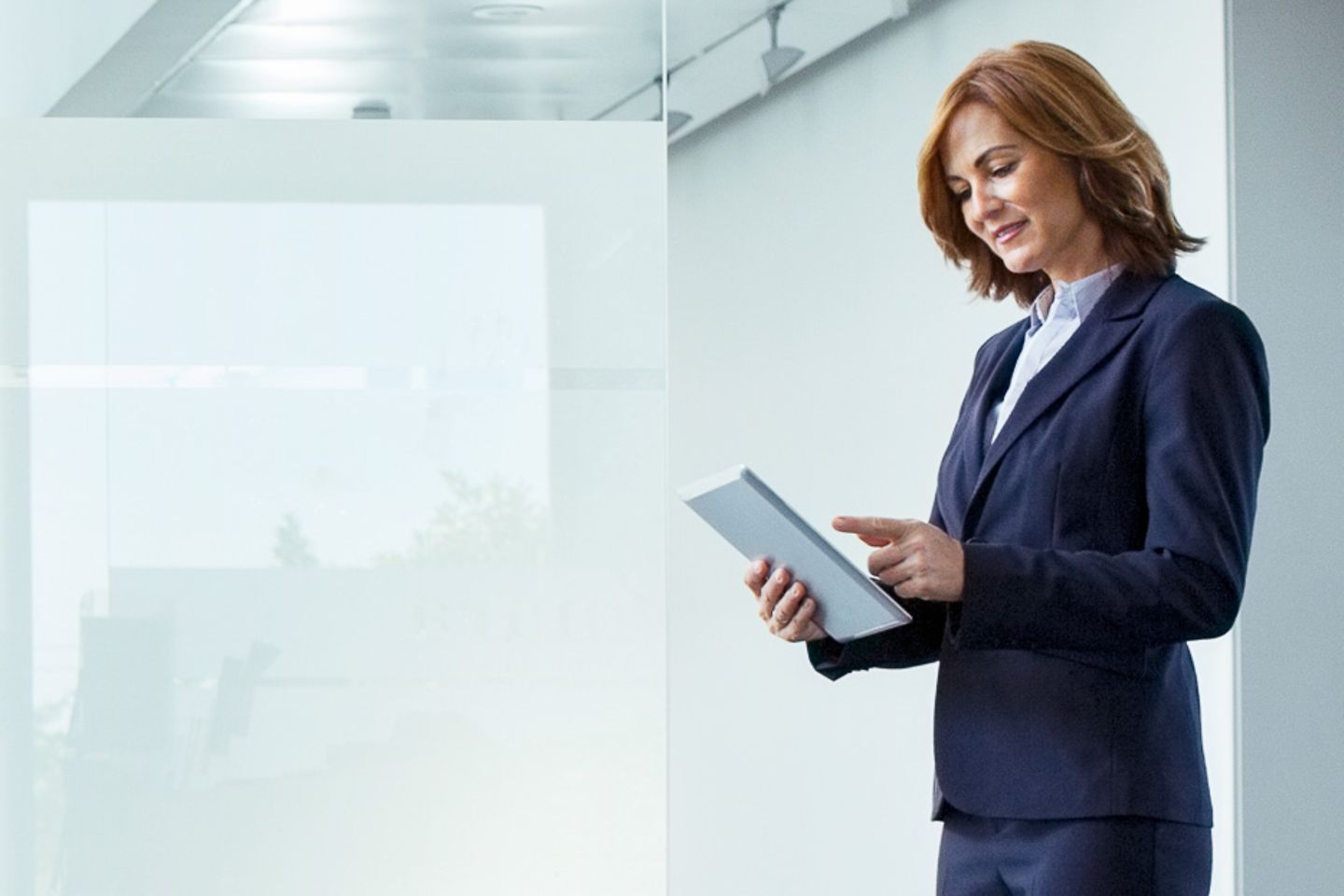 Geschäftsfrau steht vor der Fensterfront eines modernen Bürogebäudes und arbeitet auf einem Tablet