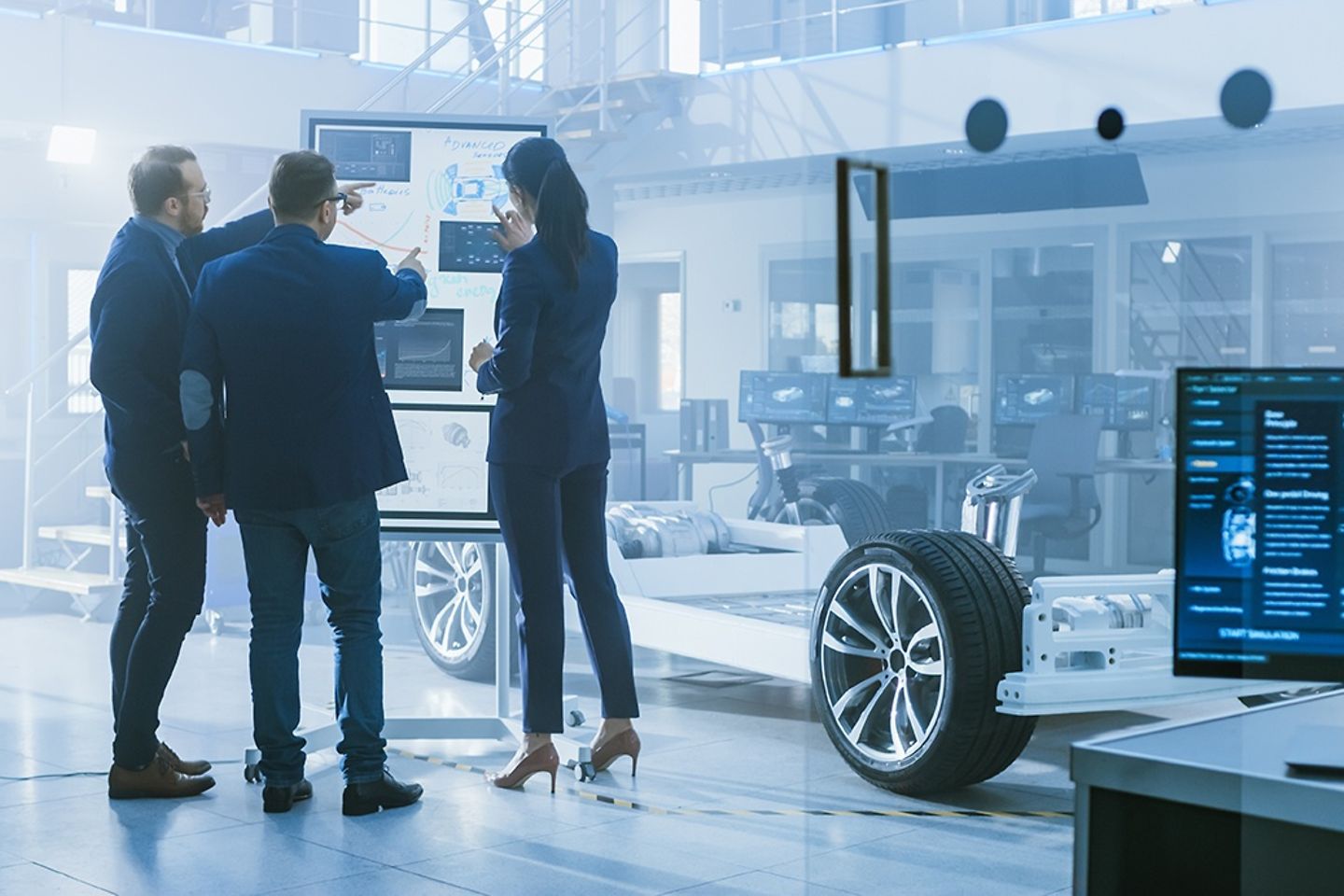 Three employees stand at a whiteboard in a factory hall with chassis.