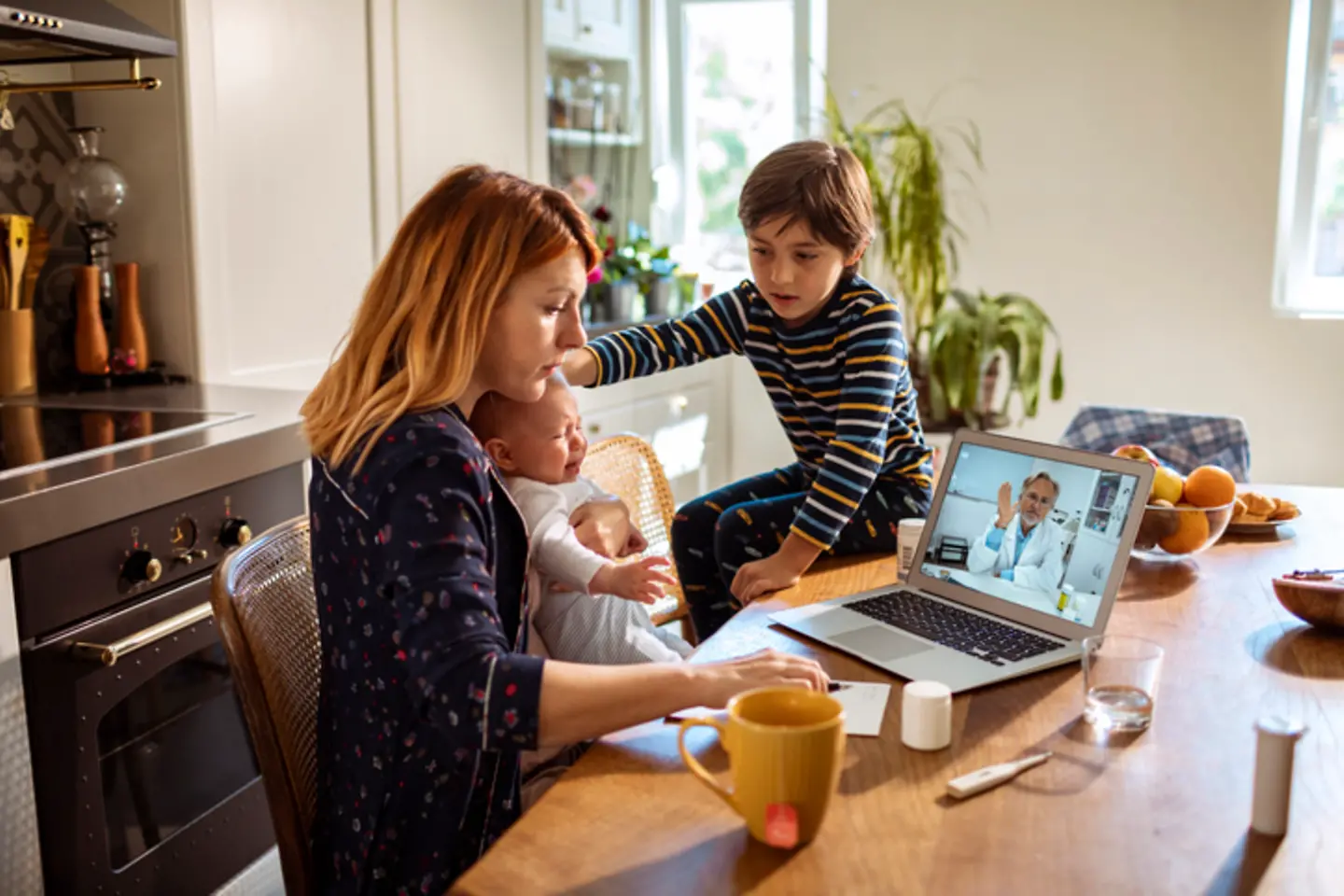 A woman sits with an infant on her lap at a table in front of a laptop, a boy is sitting on the table.