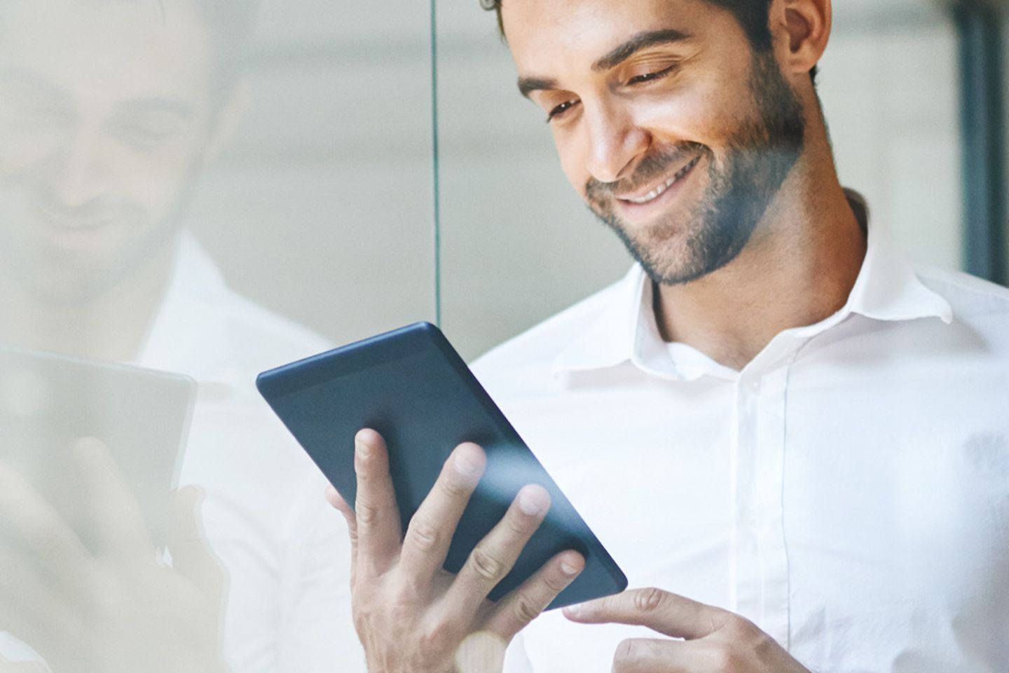 Smiling businessman leans against glass pane and looks at tablet